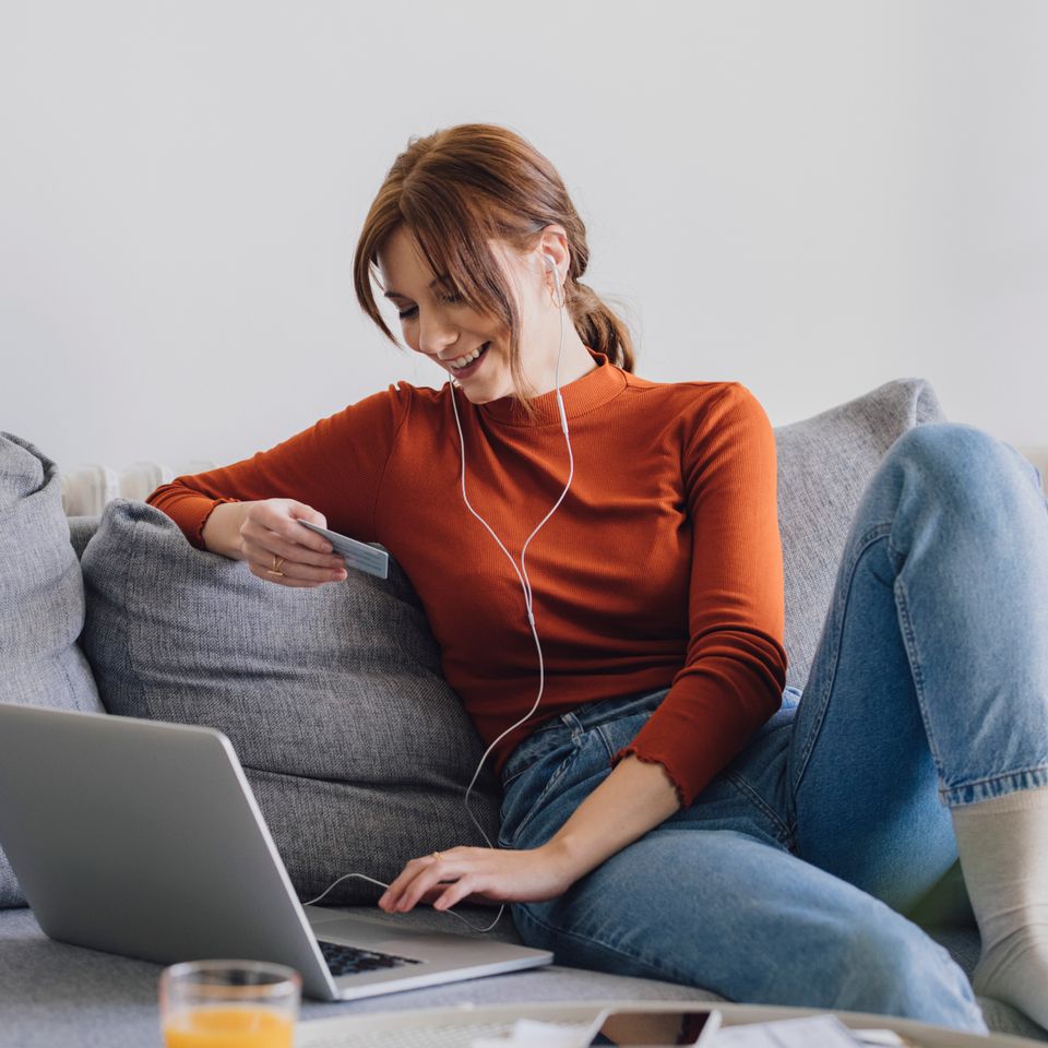 Anbieter vergleichen auf Finanzwissen.de | © iStock.com/miniseries Rothaarige Frau mit rotem Pulli sitzt auf einem Sofa und arbeitet am Laptop | © iStock.com/miniseries